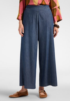 Donna che indossa pantaloni larghi in denim blu, una camicetta colorata a maniche lunghe, scarpe marroni basse e un braccialetto scuro al polso destro.