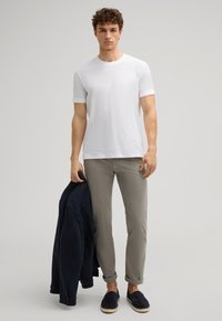 Weißes T-Shirt mit kurzen Ärmeln, hellgraue Hose, marineblauer Jacke über dem Arm, und schwarze Espadrilles. Glattes Material, lockere Passform, lässiger Stil.