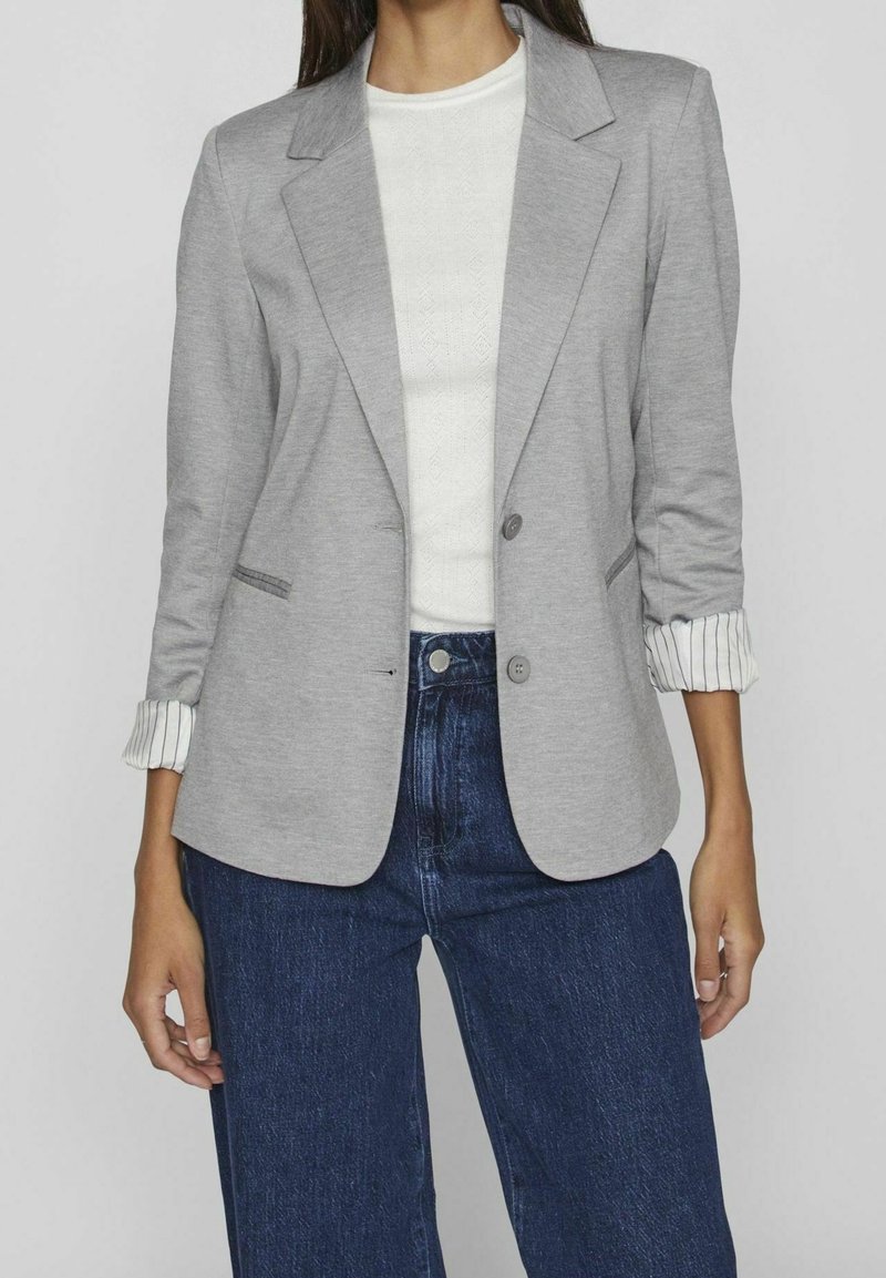 Femme portant un blazer gris clair avec des poignets rayés retroussés sur un haut blanc et un jean bleu foncé taille haute, debout, les mains détendues.