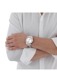 Orologio argentato con quadrante rotondo, bracciale in metallo, quadrante testurizzato argentato, lancette luminescenti e display della data. Indossato su un polso nudo in una camicia bianca.