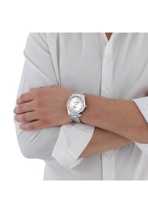 Orologio argentato con quadrante rotondo, bracciale in metallo, quadrante testurizzato argentato, lancette luminescenti e display della data. Indossato su un polso nudo in una camicia bianca.
