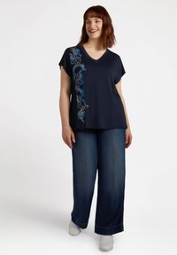 Maglietta blu navy a V con maniche corte, caratterizzata da ricami floreali blu e argentati sul lato sinistro; abbinata a jeans in denim scuro a gamba larga.
