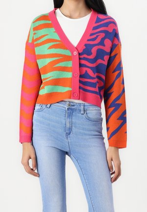 Kobieta ubrana w kolorowy kardigan na guziki z abstrakcyjnymi wzorami tygrysa i błyskawicy, nosząca biały T-shirt i jasnoniebieskie dżinsy.