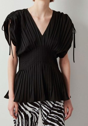 Kvinde iført en sort plisseret bluse med puffede ærmer og bukser med zebramønster, stående mod en ensfarvet lys væg.