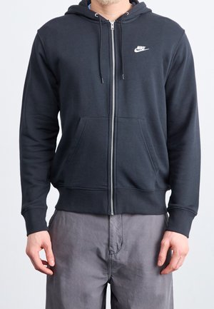 Mężczyzna w czarnej bluzie Nike z kapturem na zamek i przednimi kieszeniami oraz szarych spodniach, stojący na jednolitym jasnym tle.