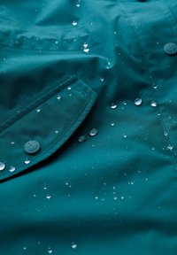 Vesitulossa teal kangas, jossa on vesipisaroita, ankkuri-designilla varustettu nappi ja näkyvä tasku, jossa on tikkaus yksityiskohtia.