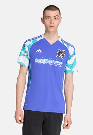 Giovane uomo che indossa una maglia da calcio blu dei Seattle Sounders con maniche a pattern geometrico e logo del team, in piedi su sfondo bianco.