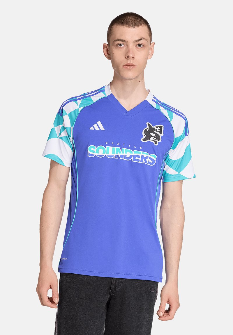 Joven vestido con una camiseta de fútbol de los Seattle Sounders de color azul, con mangas de patrón geométrico y el logo del equipo, de pie frente a un fondo blanco.