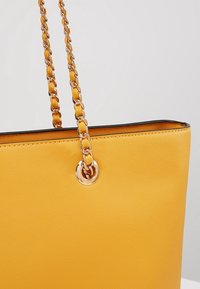 Żółta torba na ramię z teksturowaną tkaniną, uchwytami z łańcucha w kolorze złotym i okrągłym metalicznym akcentem blisko górnej krawędzi.