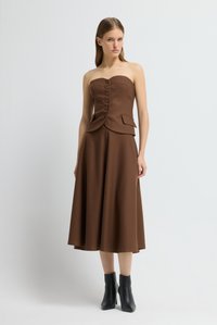 Frau in einem trägerlosen braunen Midi-Kleid mit Knöpfen an der Vorderseite und schwarzen Knöchelstiefeln, die vor einem schlichten weißen Hintergrund steht.