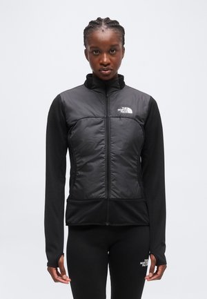 Vrouw draagt een zwart jack met rits en bijpassende leggings met The North Face-logo's, staand tegen een effen lichte achtergrond.