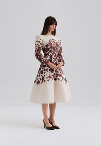 Kukallinen mekko, jossa on istuva yläosa, puffihihat ja liehuva hame. Värit sisältävät kermaisen, burgundin ja tummanruskeita yksityiskohtia.