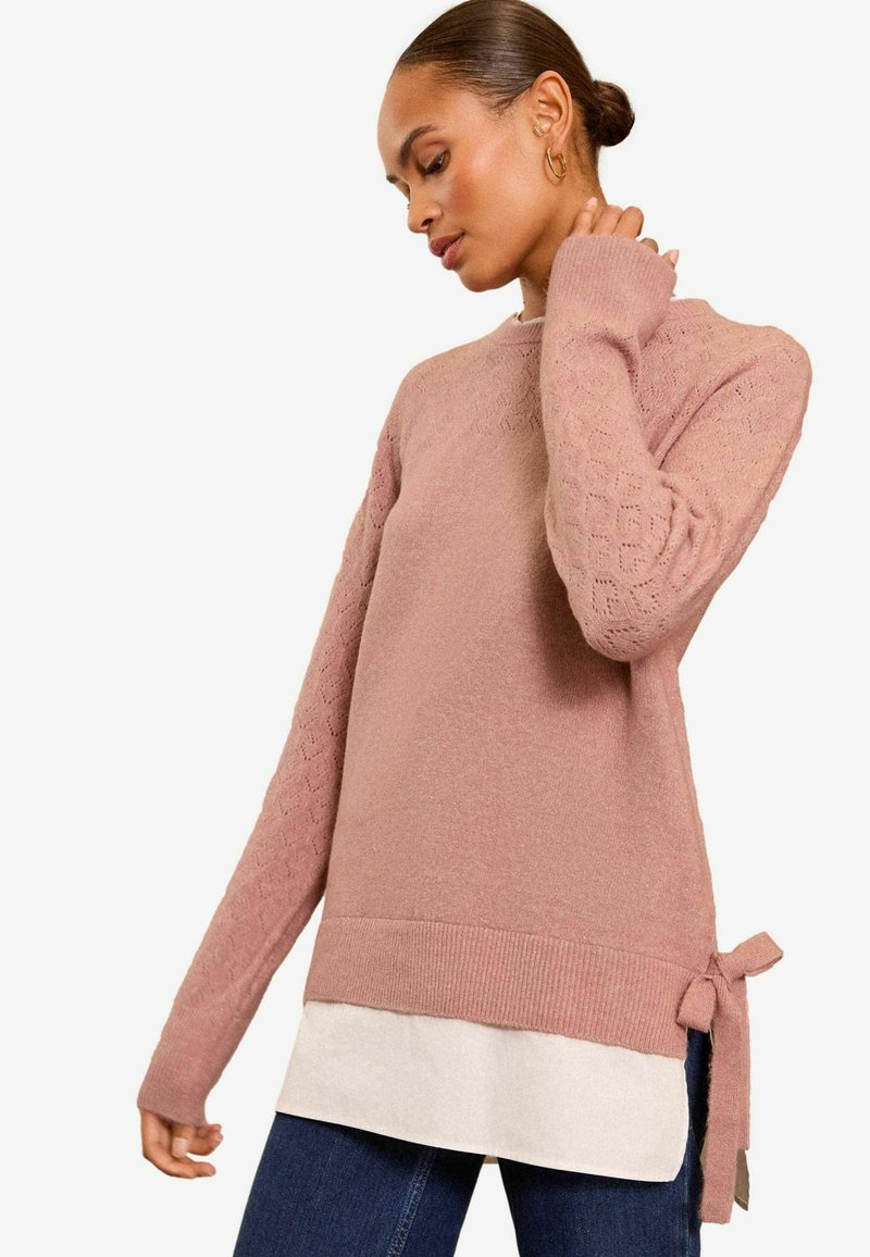 Svijetloružičasti pleteni pulover s teksturiranim rukavima i detaljem vezanja u struku, uparen s bijelim slojevitim donjim dijelom.