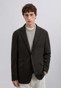 Joven con blazer verde oscuro sobre suéter gris claro y pantalones blancos, de pie con una mano en el bolsillo frente a un fondo simple.