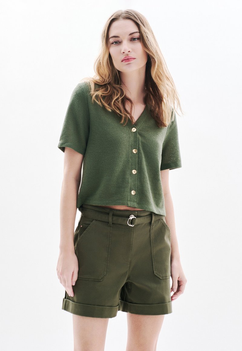 Olivgrünes kurzärmliges Blusenshirt mit V-Ausschnitt und goldenen Knöpfen, kombiniert mit olivgrünen Shorts mit Umschlägen, seitlichen Taschen und einem Gürtel.