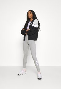 Fekete dzseki szürke ujjal, lila felső, szürke leggings fehér Nike logóval és fehér sportcipő narancssárga akcentussal. Egyszerű és alakra simuló kialakítás.