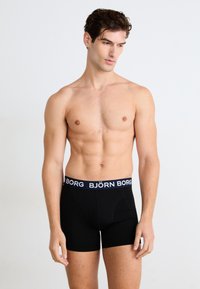 Svart boxertruser laget av elastisk stoff med ensfarget design, med en midjebånd i blått og hvitt logo. Glatt tekstur og tettsittende design.