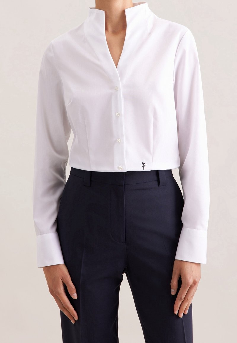 Femme portant une chemise blanche cintrée à boutons avec col montant et petite broderie florale noire près de l'ourlet, associée à un pantalon sombre.