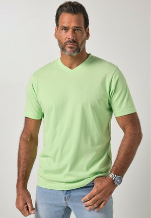 Homme aux cheveux courts et foncés avec une barbe, portant un t-shirt vert clair à manches courtes et col en V, et un jean bleu clair, debout avec une main dans la poche.