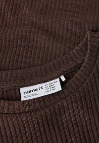 Brązowa prążkowana dzianinowa bluzka z zaokrąglonym dekoltem. Metka wskazuje rozmiary EU 122/128, US 6X/7, CA 7-8A, UK 7-8Y. Wyprodukowano w Chinach.