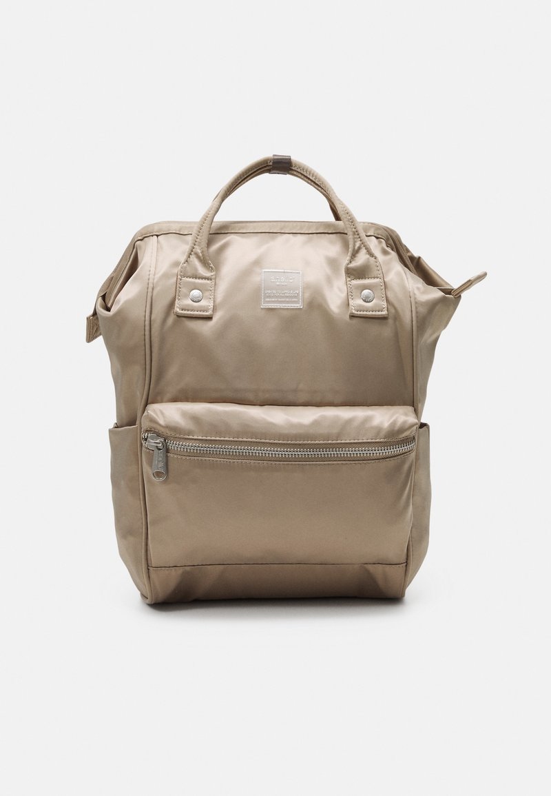 Sac à dos en nylon beige avec poignées supérieures, poche zippée frontale, poches latérales et une petite étiquette carrée au centre avant.