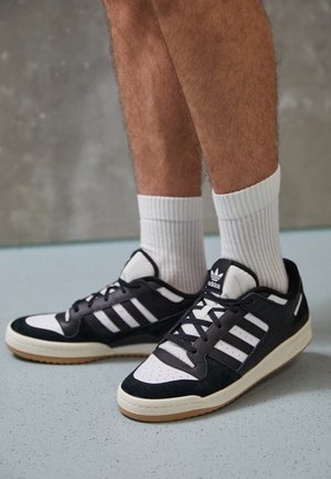 Person iført sorte og hvide Adidas-sneakers med hvide ribstrikkede sokker, der står på en lyseblå plettet gulv.