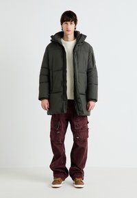 Polstret olivengrøn parka med hætte og frontlynlås, parret med burgunderfarvede corduroy cargo-bukser med lommer og trykknapper. Brune sko.