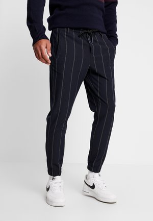 Mężczyzna w czarnych dresowych spodniach w prążki i białych sneakersach Nike, stojący z rękami w kieszeniach na jednolitym tle.