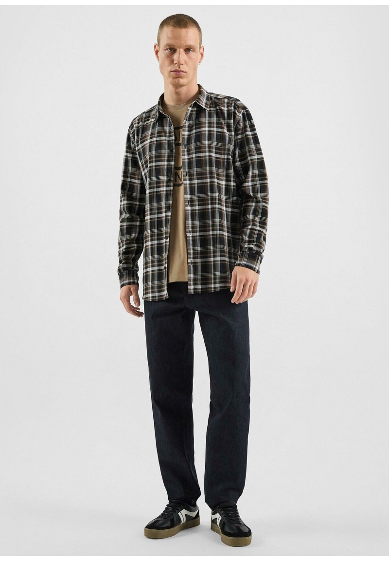 Camicia a scacchi nera e marrone con maniche lunghe e chiusura a bottoni, indossata sopra una t-shirt grafica beige, abbinata a jeans scuri e sneakers.