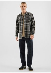 Schwarz-braun kariertes Hemd mit langen Ärmeln und Knopfleiste, über einem beigen Grafik-T-Shirt getragen, kombiniert mit einer dunklen Jeans und Sneakers.