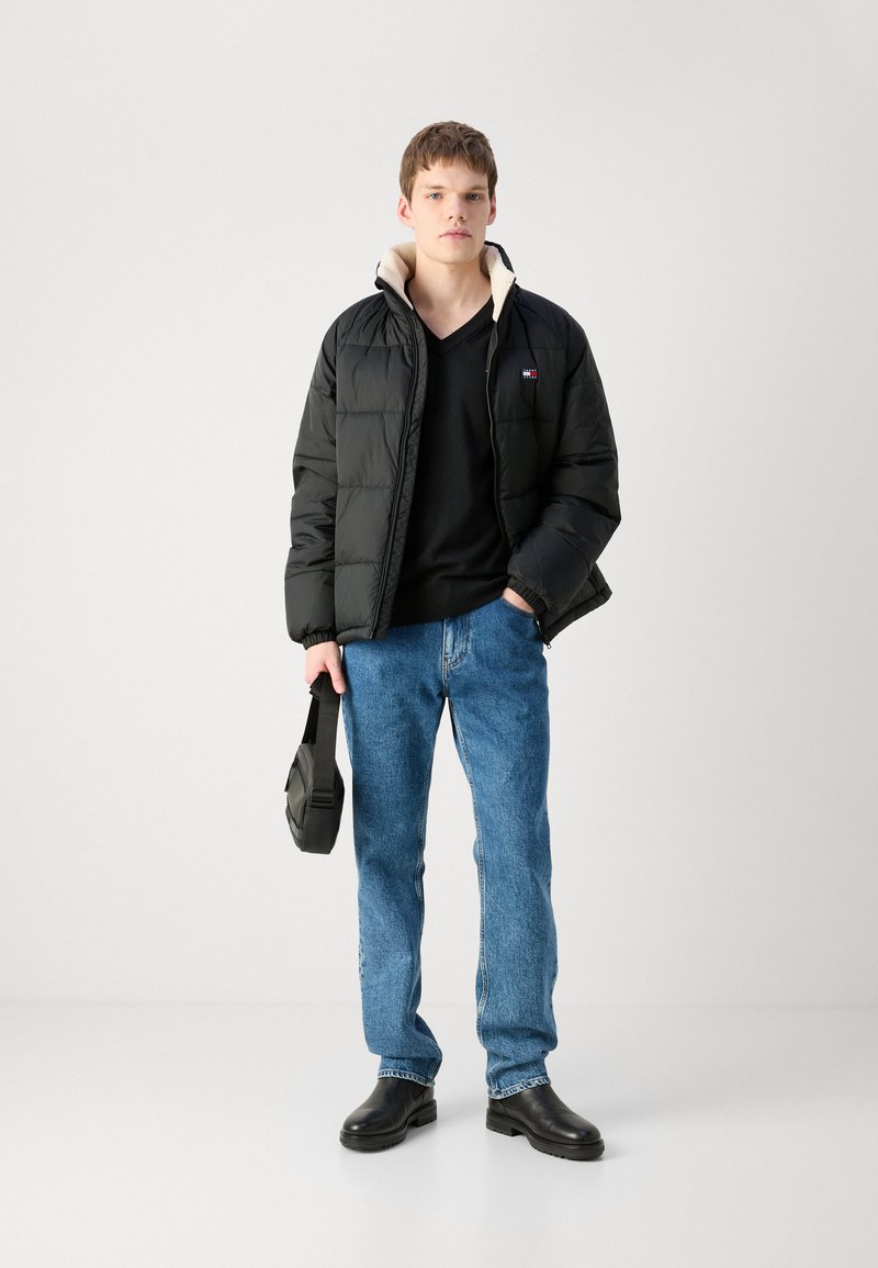 Schwarze Daunenjacke mit beiger Innenausstattung, schwarzer V-Ausschnitt-Pullover, blaue Jeans und schwarze Stiefeletten. Hält eine kleine schwarze Tasche.