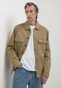 Un homme portant une veste beige à boutons sur un t-shirt blanc et un jean bleu, se tenant devant un fond gris clair uni.