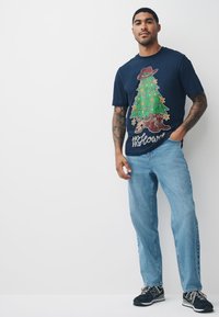 Tamsiai mėlynas medvilninis marškinėlis su spalvotu Kalėdų eglutės ir batų piešiniu, derinamas su šviesiai mėlynomis džinsinėmis kelnėmis ir juodais sportbačiais.