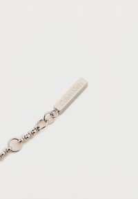 Sidabrinis metalinis apyrankės, kuriame yra ovalūs žiedai ir tiesioginė etiketė su užrašu "Calvin Klein". Lygi tekstūra ir minimalistinis dizainas.