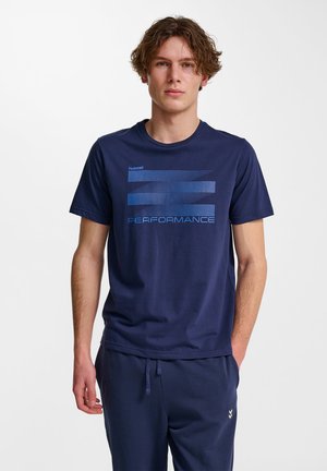 Jonge man die een donkerblauw T-shirt met blauwe tekst "PERFORMANCE" en een bijpassende donkerblauwe broek draagt, staand tegen een witte achtergrond.