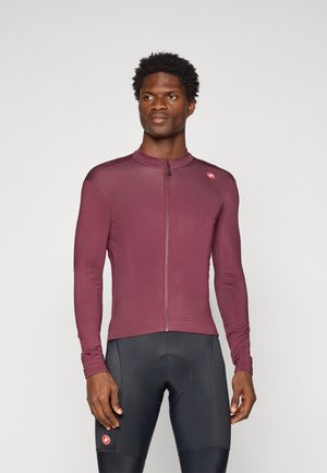 Mann in einem maroonfarbenen, gepolsterten Radtrikot mit Reißverschluss und schwarzen gepolsterten Radhosen, steht vor einem einfachen hellen Hintergrund.