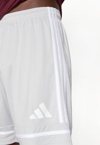 Weiße Sportshorts mit Adidas-Logo und drei Streifen an der Seite, getragen von einer Person mit dunkler Hautfarbe und einem dunkelroten Oberteil.