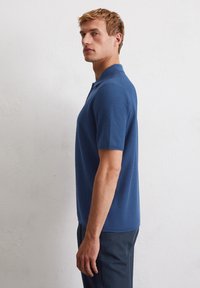Polo shirt in diepblauw, korte mouwen, textuurstof en kraag. Draagt bij donkere broeken, gepresenteerd tegen een neutrale achtergrond.