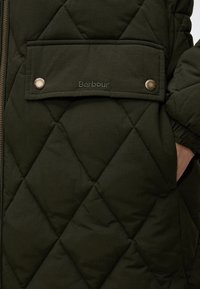 Poche de veste matelassée vert foncé avec logo Barbour et deux boutons-pression dorés, montrant partiellement une main à l'intérieur de la poche latérale.