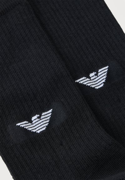 Paire de chaussettes noires côtelées avec logos d'aigle à rayures blanches près de la zone des orteils sur un fond clair.