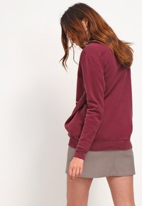 Sudadera burdeos hecha de tela suave, con un bolsillo frontal y puños de canalé, combinada con una minifalda gris claro.