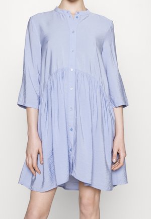 Femme portant une robe ample bleu clair à boutons, manches trois-quarts, avec un tissu froncé sous la poitrine.