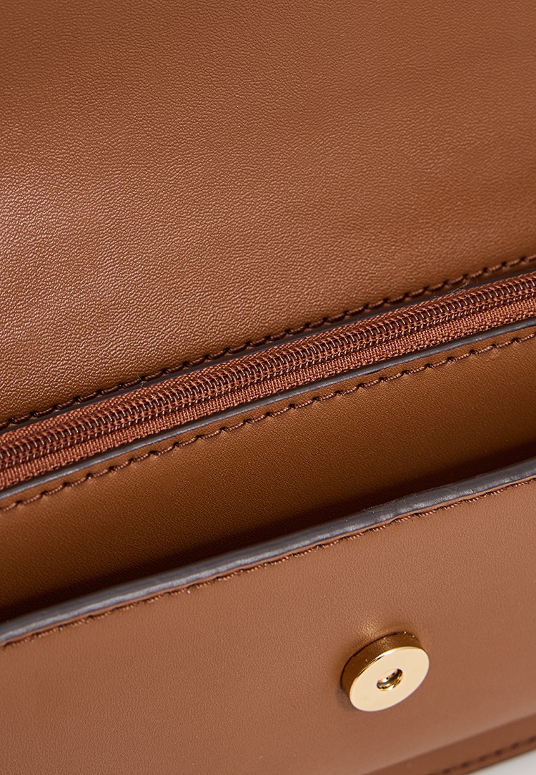 Détail d'un sac en cuir brun avec un fermoir à pression doré et un compartiment zippé, mettant en valeur une texture lisse et des lignes épurées.