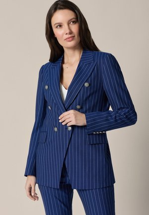 Kobieta ubrana w granatowy, dwurzędowy garnitur w prążki z srebrnymi guzikami, pozująca z jedną ręką przy zapięciu marynarki.
