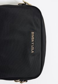 Sort tekstilpose med en glat overflade, guldtonet logo "BIMBA Y LOLA" og en lynlåslukning. Detaljerne inkluderer afrundede kanter og guldhardware.