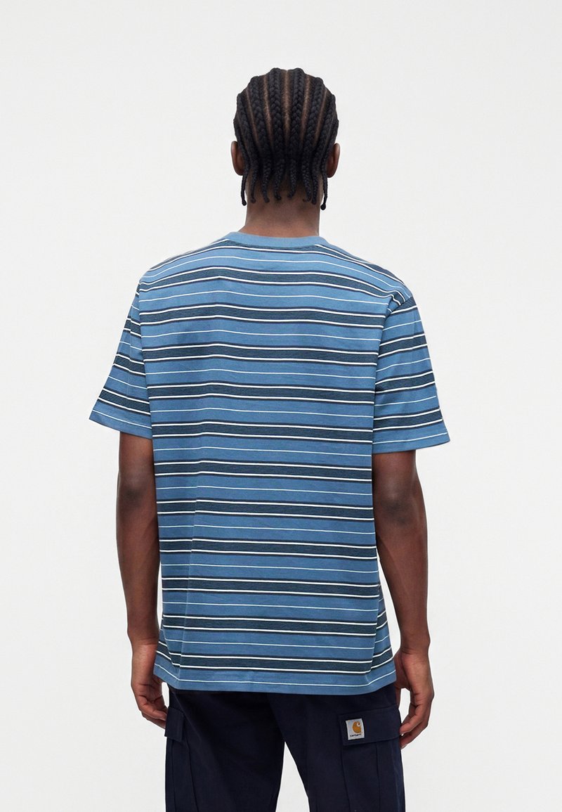 Mann mit Cornrow-Zöpfen, der ein blau gestreiftes kurzärmeliges Hemd und dunkle Cargohosen trägt, mit dem Rücken zu einem einfarbigen Hintergrund steht.