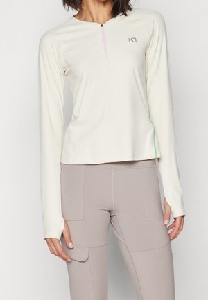 Mujer con top crema de manga larga con cremallera de cuarto y pantalones ajustados color topo con detalles de bolsillo, de pie contra un fondo liso.