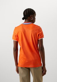 Orange polo-skjorta med en texturerad design och små perforeringar, vita detaljer på ärmarna och en flerfärgad krage.
