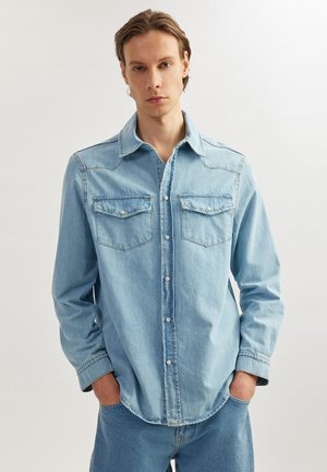 Camisa de mezclilla azul claro con cuello clásico, dos bolsillos en el pecho y cierre de botones. Mangas largas con puños abotonados y dobladillo curvado.