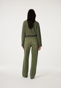 Vrouw met lang, donker haar staat met haar rug naar de kijker, gekleed in een olijfgroene trui en bijpassende hoge wijdbenige broek.